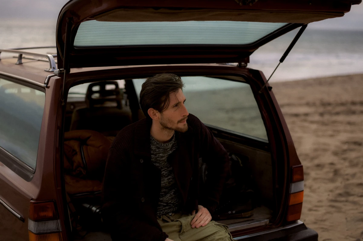 Young man sitting in open tailgate of station wagon by the beach for ERGO road trip campaign